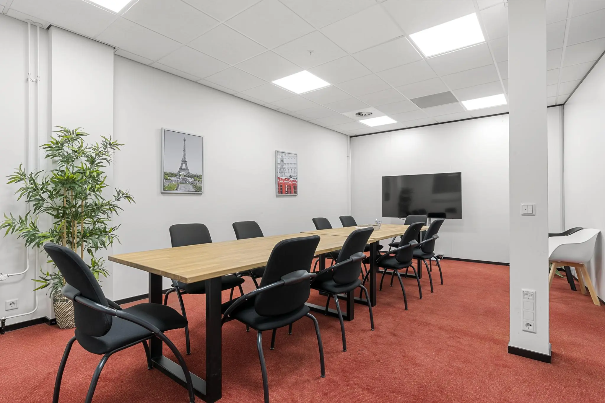 Vergaderruimte met lange houten tafel, zwarte stoelen, rode vloerbedekking en een flatscreen aan de muur.